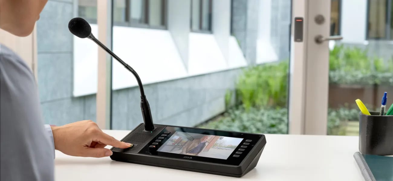 A hand pressing the talk button on a desktop paging console with a gooseneck microphone.